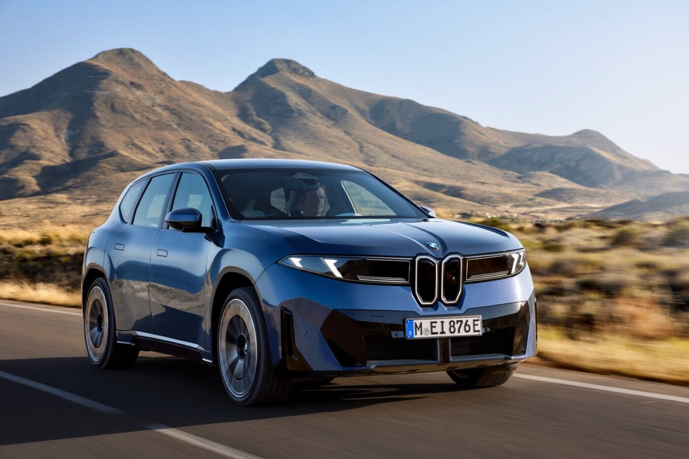 BMW iX3 M60 xDrive elektrikli SUV’nin dinamik bir sürüş sahnesi, yol kenarında hızlı şarj istasyonu