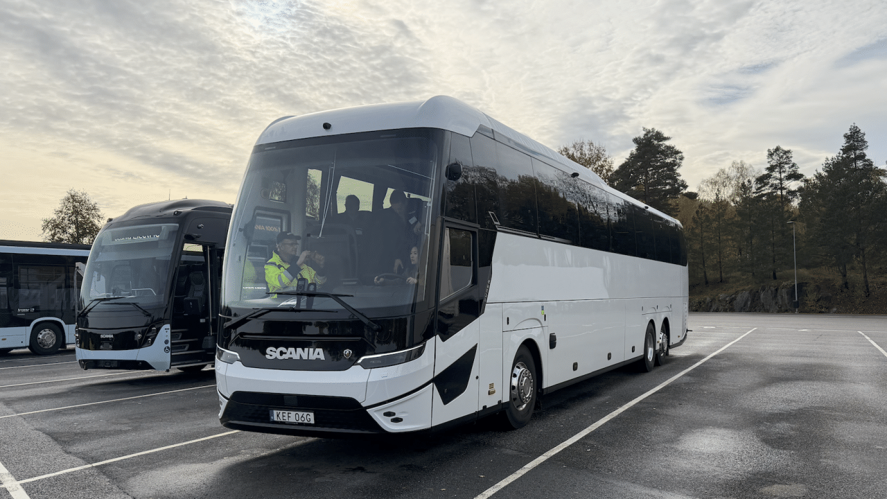 Yeni Scania Touring elektrikli otobüsü, şehirlerarası bir yolda ilerliyor, akşam ışığı altında. Otobüs mavi ve gümüş renklerde, alçak açıdan çekilmiş.
