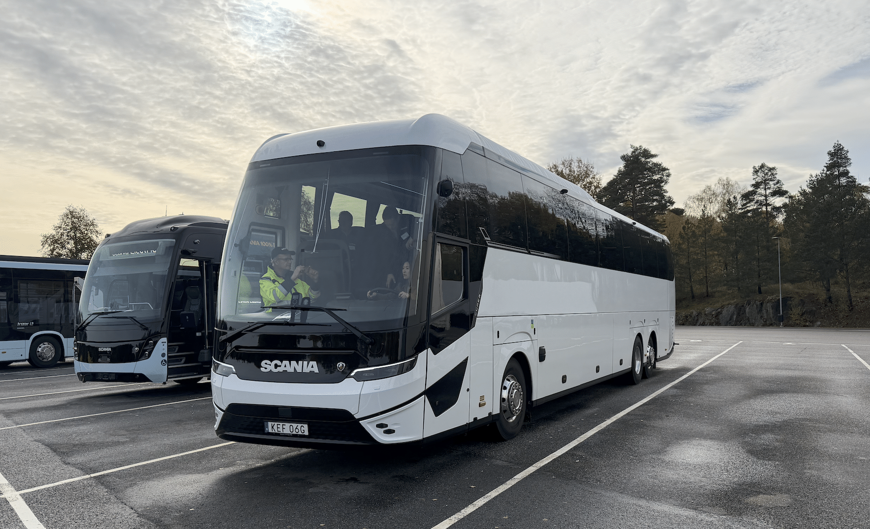 Yeni Scania Touring elektrikli otobüsü, şehirlerarası bir yolda ilerliyor, akşam ışığı altında. Otobüs mavi ve gümüş renklerde, alçak açıdan çekilmiş.