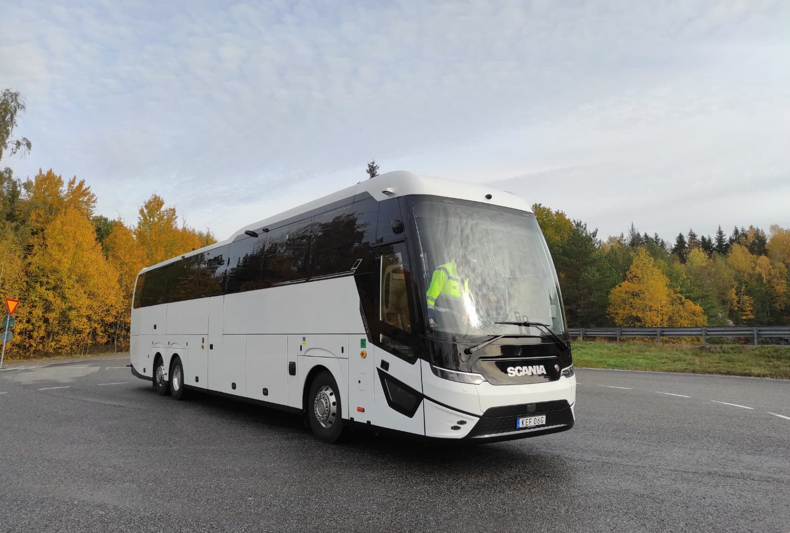 Yeni Scania Touring elektrikli otobüsü, şehirlerarası bir yolda ilerliyor, akşam ışığı altında. Otobüs mavi ve gümüş renklerde, alçak açıdan çekilmiş.