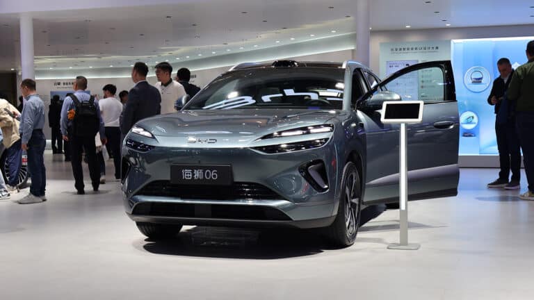 Bir Avrupa şehrinde şarj istasyonunda şarj olan modern bir elektrikli otomobil, arka planda uluslararası ticareti simgeleyen hareketli bir liman ve yük gemileri.