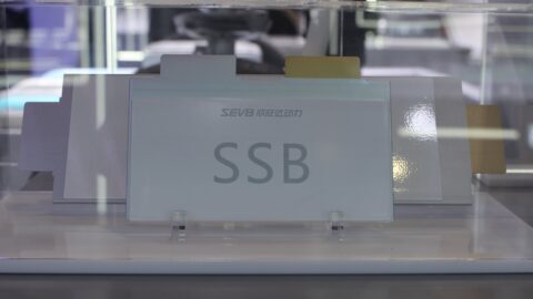 Bir Avrupa şehrinde şarj istasyonunda şarj olan modern bir elektrikli otomobil, arka planda uluslararası ticareti simgeleyen hareketli bir liman ve yük gemileri.