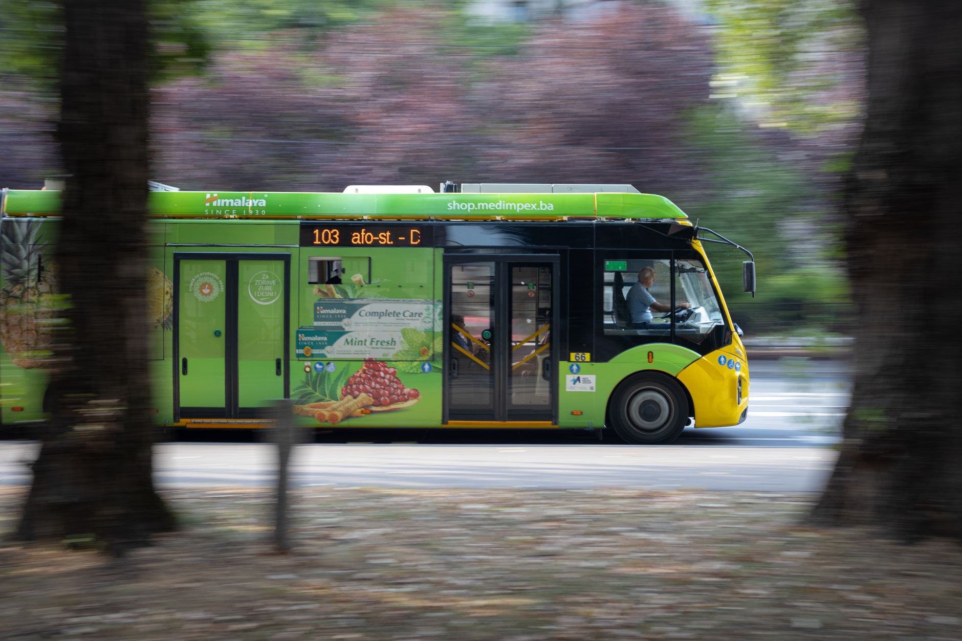 Elektrikli bir şehir içi otobüs, modern bir otobüs durağının önünde yolcu alıyor. Arka planda ağaçlar ve mavi gökyüzü var. Görselde yazı, logo veya marka bulunmuyor.