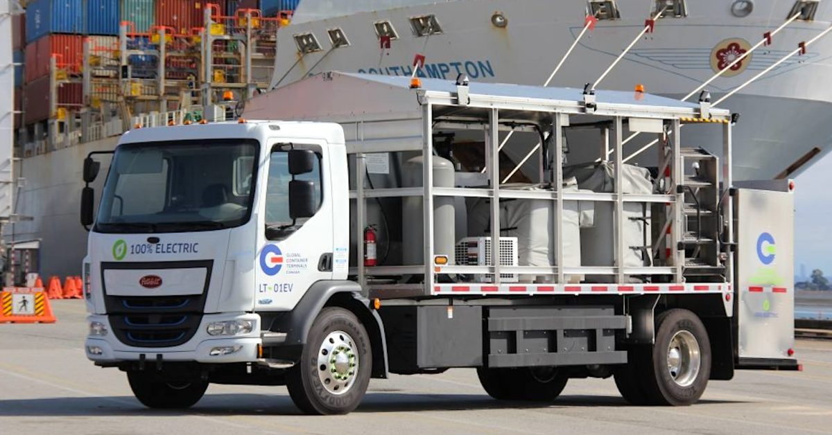 Elektrikli Peterbilt tanker kamyonu Vancouver Limanı'nda şarj olurken, arka planda konteyner yığınları ve vinçler görülüyor.