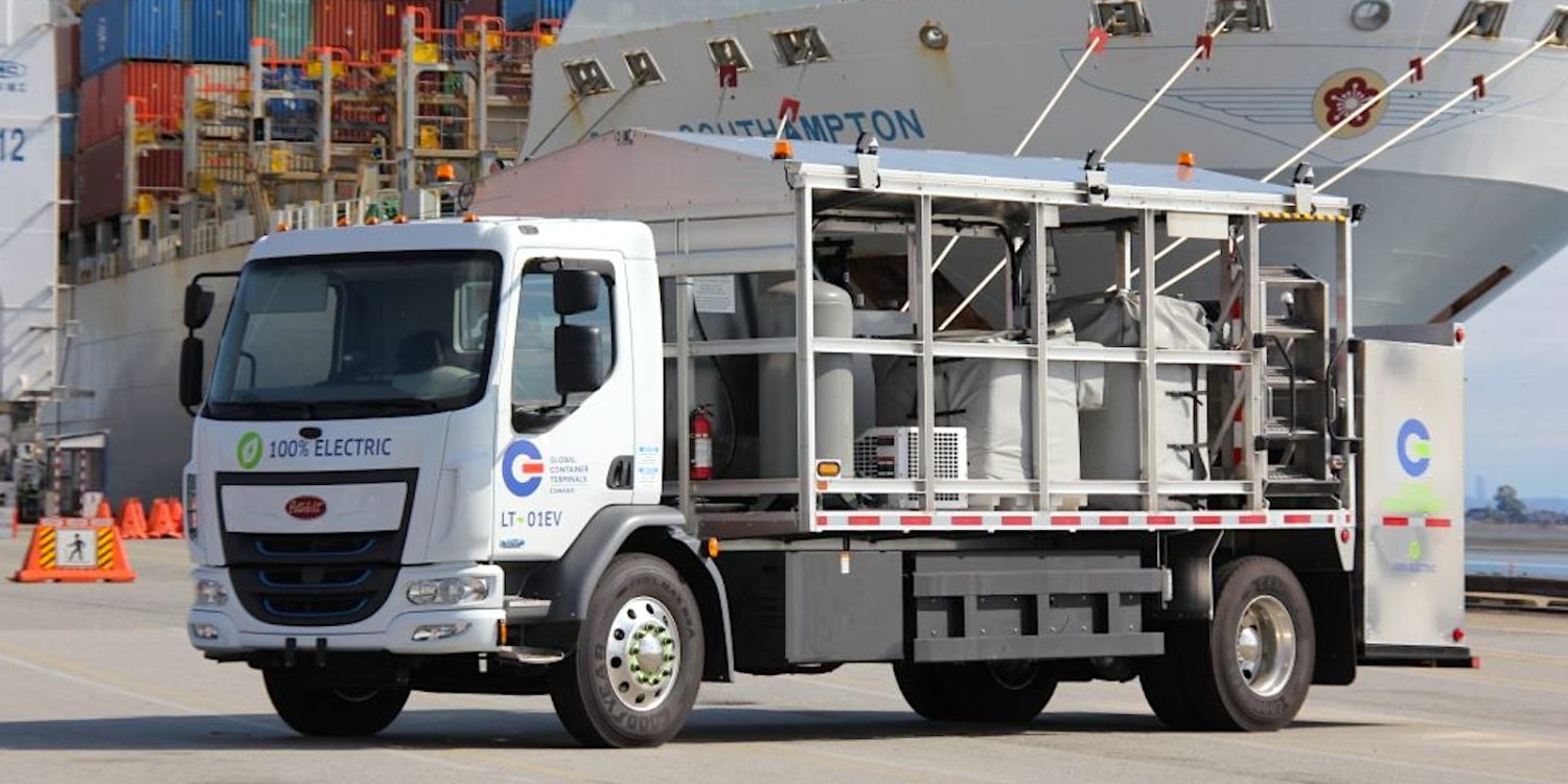 Elektrikli Peterbilt tanker kamyonu Vancouver Limanı'nda şarj olurken, arka planda konteyner yığınları ve vinçler görülüyor.