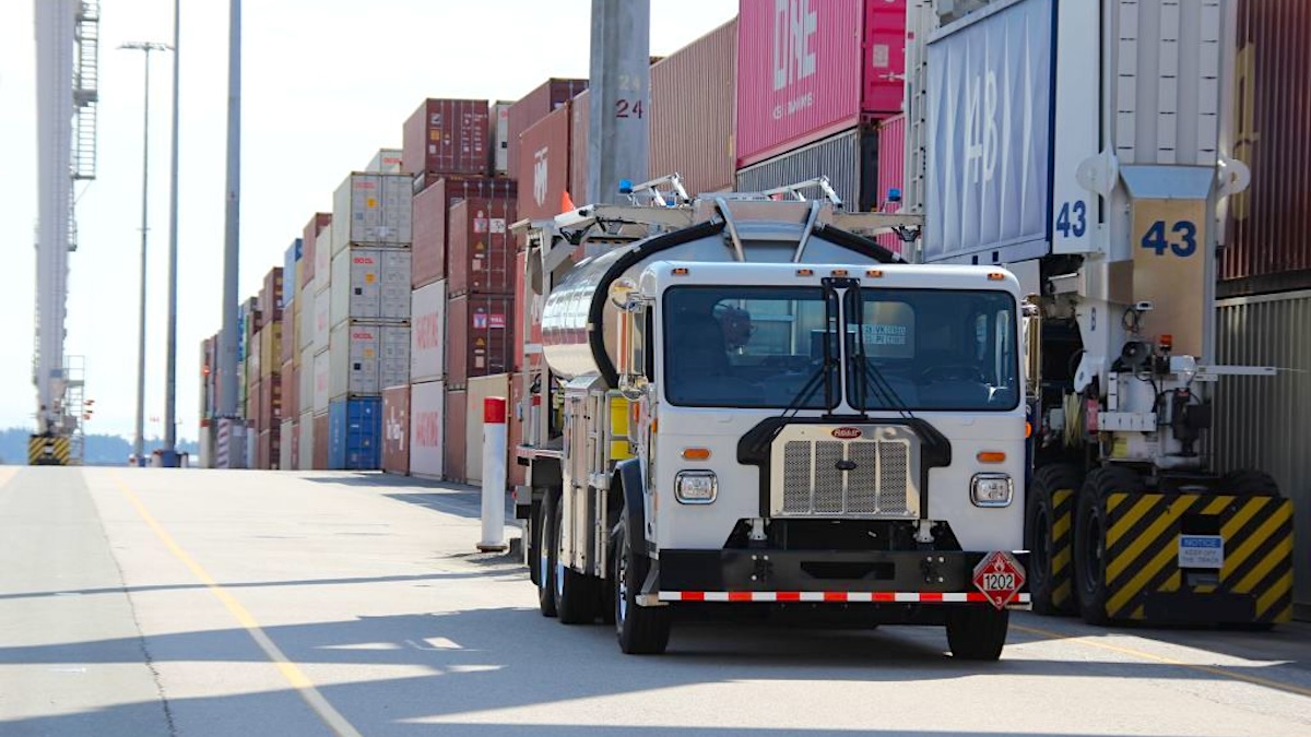 Elektrikli Peterbilt tanker kamyonu Vancouver Limanı'nda şarj olurken, arka planda konteyner yığınları ve vinçler görülüyor.
